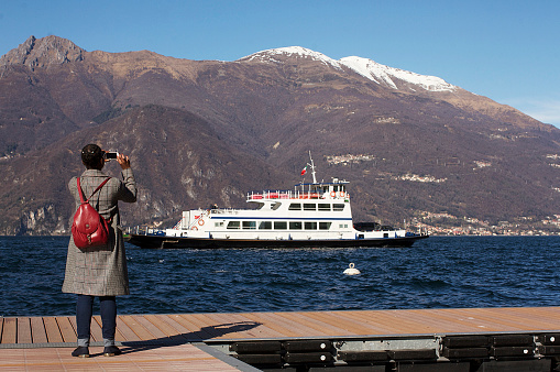 Colour photograph of a young woman taking a photo of a boat & mountains in Verenna, Italy