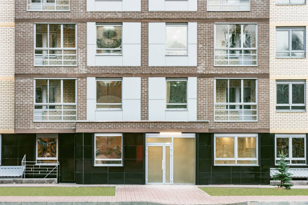 A contemporary apartment building featuring red brick and light-colored walls with multiple metal-railed balconies. An autumn tree in the foreground adds organic texture and contrast to the clean architectural lines. The scene conveys a calm, urban residential atmosphere with a mix of modern design elements.