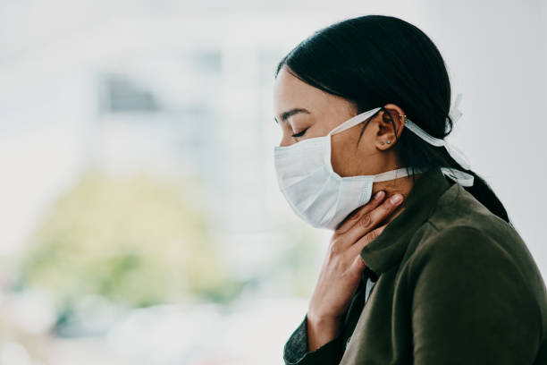 Take care, Covid-19 is here Shot of a young woman wearing a mask and suffering from throat pain in a doctor’s office symptom stock pictures, royalty-free photos & images