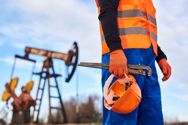 Oil worker in orange uniform and helmet on of background the pump jack and blue sky. Cropped close-up snapshot of a man wearing blue overalls orange vest and gloves, holding a pipe wrench and a helmet on foreground, oil pump jack on background. Concept of petroleum industry. oil field stock pictures, royalty-free photos & images