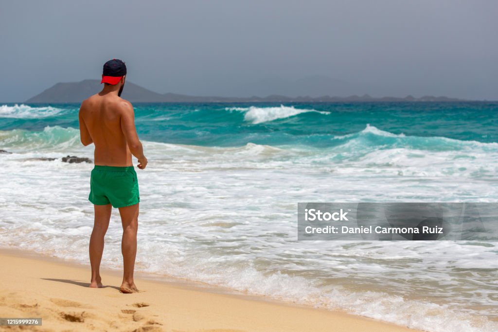 A young man with green swimming trunks walking stands on the sand while looking at a beach of Fuerteventura, Canary Islands, Spain A young handsome man standing on the sand in the beach. Canary Islands One Young Man Only Stock Photo