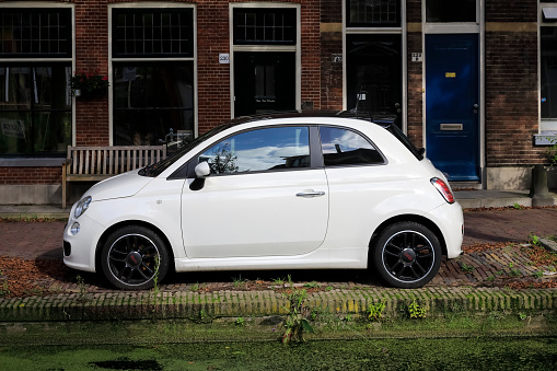 Delft, The Netherlands - October 10, 2019: A car, Fiat 500 in the narrow street parked between brick buildings and a bank of inland waterway. This is a car by Italian manufacturer, on sale since 2007.
