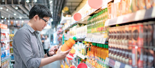 Asian man shopping orange juice using phone Asian man choosing orange juice in supermarket using smartphone to check shopping list. Male shopper with shopping cart selecting beverage bottle product in grocery store. megastore stock pictures, royalty-free photos & images