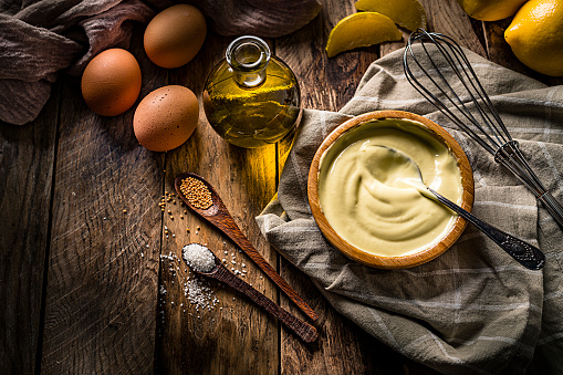 Food recipe: top view homemade mayonnaise in a wood bowl shot on rustic wooden table. Ingredients for preparing homemade mayonnaise like olive oil, eggs, lemon and salt are around the prepared mayonnaise bowl. Predominant colors are brown and yellow. High resolution 42Mp studio digital capture taken with SONY A7rII and Zeiss Batis 40mm F2.0 CF lens