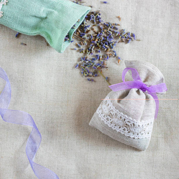 istock Small linen sack filled with dried lavender decorated with lacework and violet ribbon coqueand and one sachet is opened. Top view on thenatural flaxen background. Square with copy space 1216008490