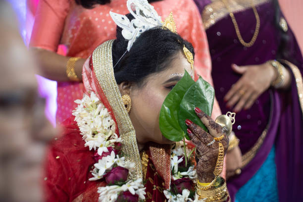 colpo di fuoco selettivo della sposa bengalese in saree rossa e gioielli nascosti dietro una foglia di betel paan mentre la sua famiglia guarda - image title foto e immagini stock