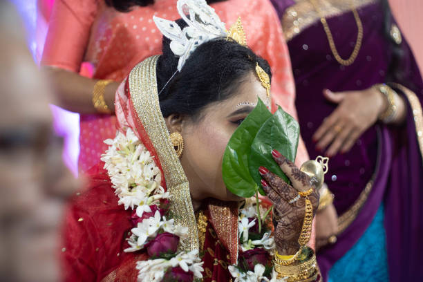 tiro de foco seletivo de noiva bengali em saree vermelho e jóias escondidas atrás de uma folha de betel paan enquanto sua família olha em - image title - fotografias e filmes do acervo