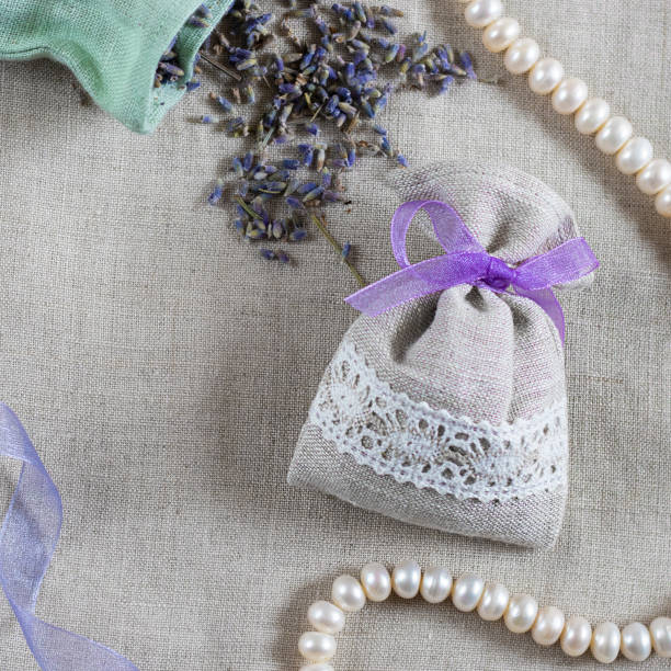 istock Small linen sack filled with dried lavender decorated with lacework and violet ribbon, two sachets, one is opened and pearl necklace. Top view on thenatural flaxen background. Square 1216009994