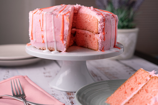 A pink buttercream frosted cake with red and white glaze dripping down the sides. The cake sits on an elevated white cake stand. Pink napkins and forks on the table. A slice of the cake nearby.