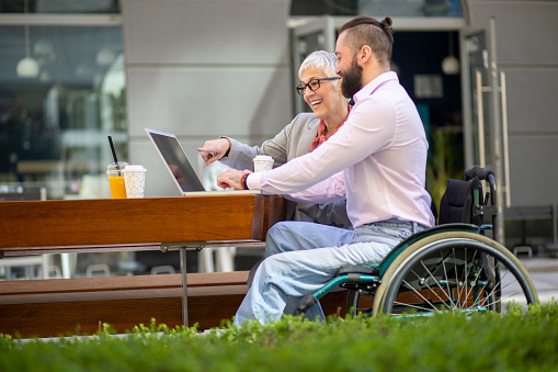 Disability smiling  man with beautiful senior mature grey hair woman on meeting out of office. They are sitting in a park on a bench, drinking coffee and finishing work on a laptop online. He is motionless and sitting in a wheelchair.