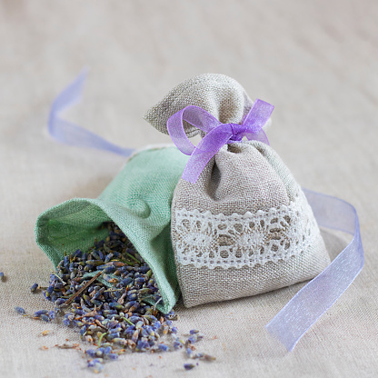Small linen bag filled with dried lavender decorated with lacework and violet ribbon coque, two sachets, one is opened. Square. Composition on thenatural flaxen background.