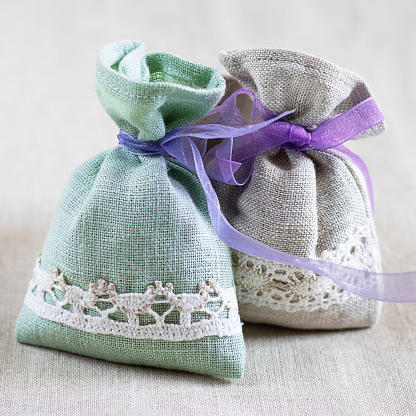 Two small linen sacks filled with dried lavender decorated with lacework and violet ribbon coque, aroma sachet. Square. Closeup composition on the natural flaxen background.