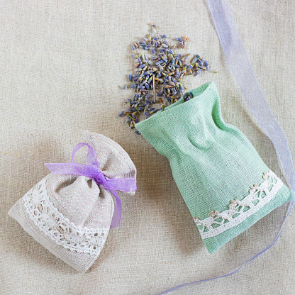 Small linen bag filled with dried lavender decorated with lacework and violet ribbon coque, two sachets, one is opened. Square with copy space. Top view on thenatural flaxen background.