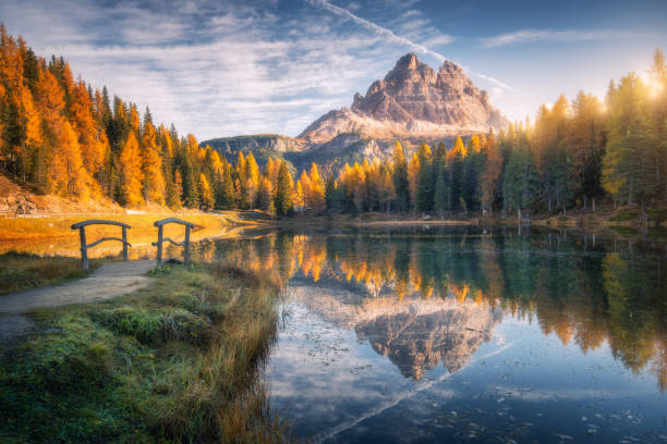 Photo of Lake with reflection in mountains at sunrise in autumn in Dolomites, Italy. Landscape with Antorno lake, small wooden bridge, trees with orange leaves, high rocks, blue sky in fall. Colorful forest