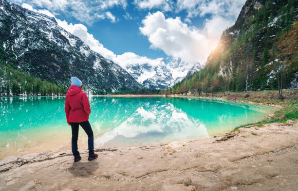 la giovane donna in giacca rossa è in piedi sulla riva del lago con acqua azzurra nella giornata di sole in primavera. paesaggio con ragazza, riflessione in acqua, montagne innevate, cielo blu con nuvole, alberi verdi. viaggio - spring forest scenics reflection foto e immagini stock