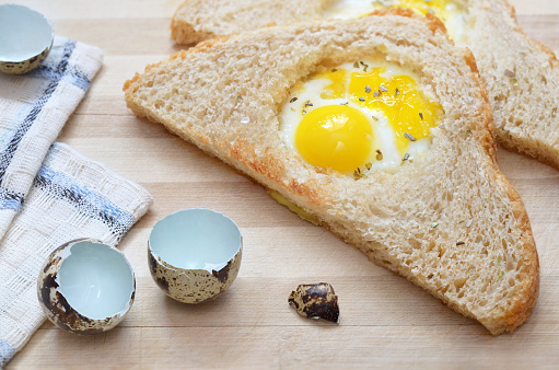 Fried slice of bread with quail egg on a wooden cutting board with a kitchen towel and eggshells, selective focus. Healthy eating concept.