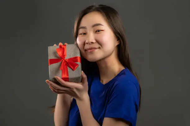 Excited asian girl received gift box with bow. She is happy and flattered by attention. Korean girl on grey background. Studio portrait.