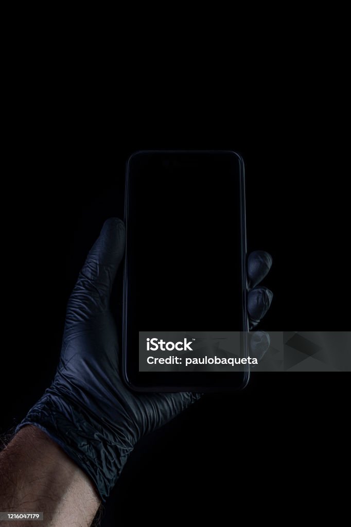 Hand holding a black phone isolated on a black background with a black gloves. Hand holding a black phone isolated on a black background with a black gloves. Desinfection against coronavirus. Black Background Stock Photo