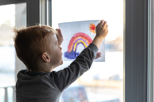Young Boy sticking his drawing on home window during the Coronavirus Covid-19 crisis, view from inside the house. Many people are putting a rainbow to tell neighbours that people inside this house are ok. #Stayathome #cavabienaller