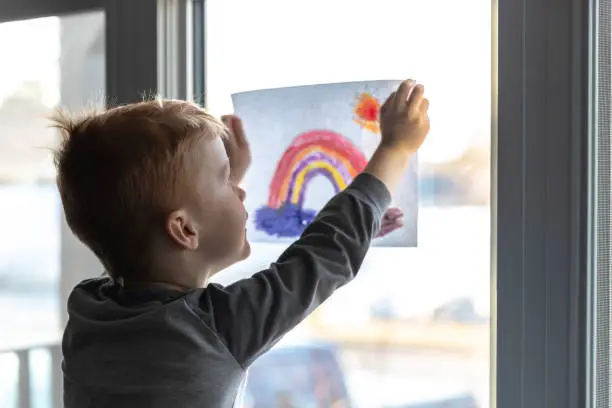 Photo of Young Boy sticking his drawing on home window during the Covid-19 crisis