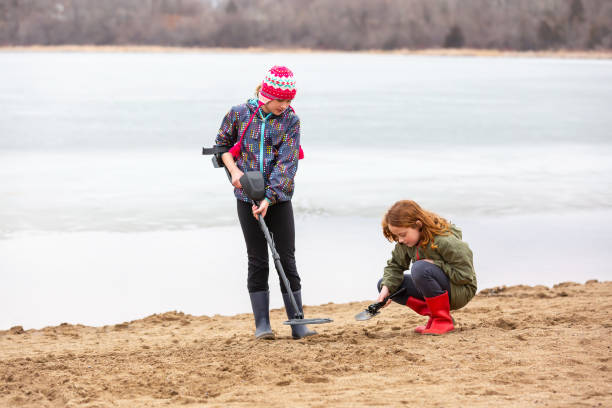 Two young girls (sisters) working together to look for treasures in the sand at the beach on an early springtime day. There is still some ice on the lake behind them. The older sister is holding the metal detector while the younger sister holds a small shovel, ready to dig for treasure.