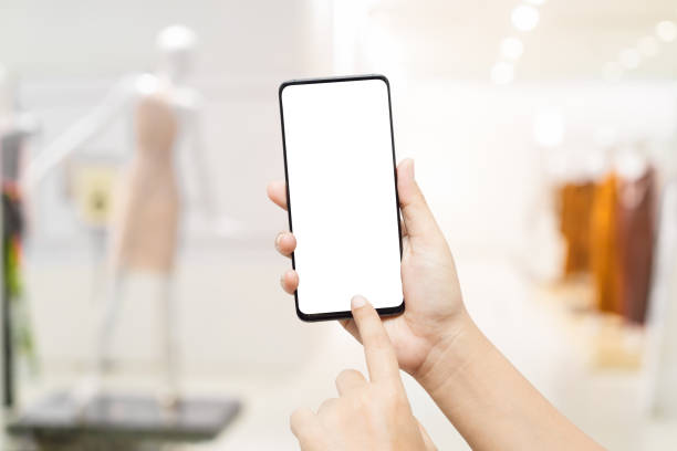 istock Mockup cellphone image hand holding smart phone with blank white screen for your advertisement with blurred abstract background of bright women clothing fashion store. Mock up mobile phone. 1216081885