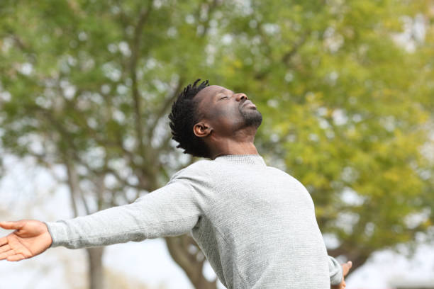 Black man breathing fresh air stretching arms in a park Black man breathing fresh air stretching arms in a park with a green tree in the background arms outstretched stock pictures, royalty-free photos & images