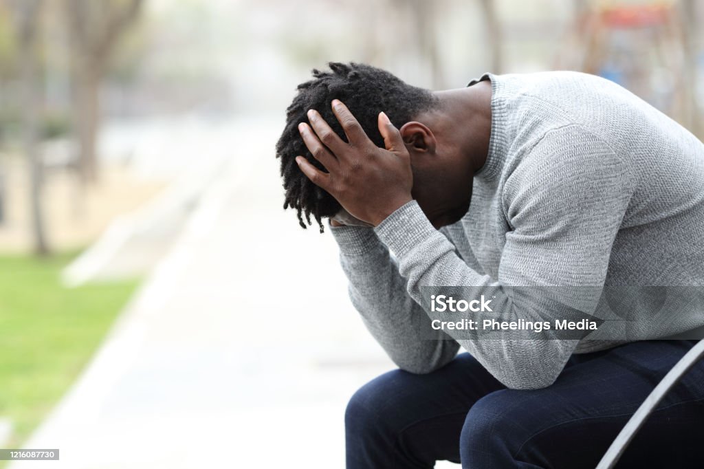 Sad depressed black man on a bench in a park Side view portrait of a sad depressed black man sitting on a bench in a park Men Stock Photo