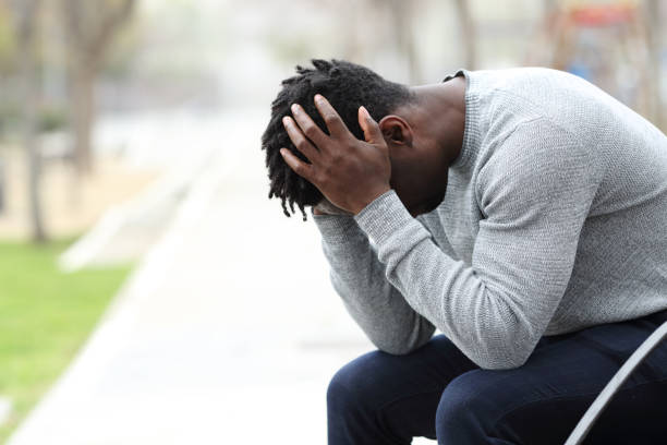 Sad depressed black man on a bench in a park Side view portrait of a sad depressed black man sitting on a bench in a park man regret stock pictures, royalty-free photos & images