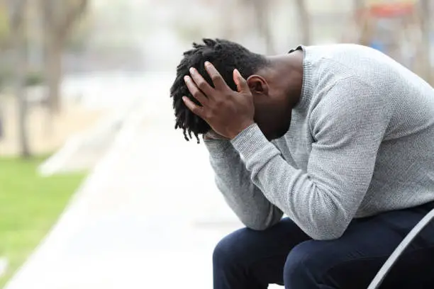 Side view portrait of a sad depressed black man sitting on a bench in a park
