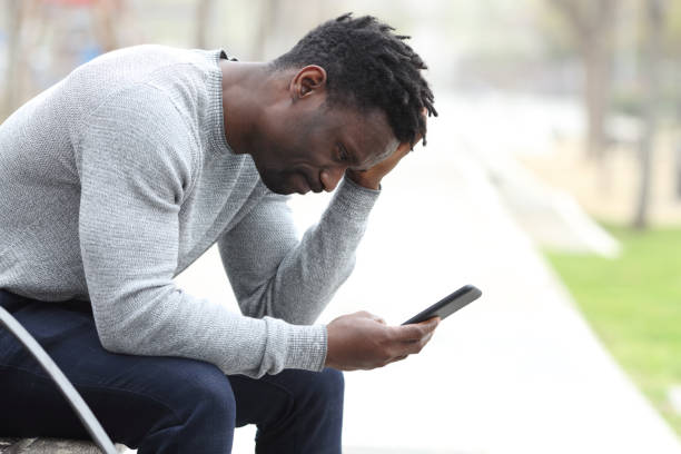 Sad black man complaining checking mobile phone Side view portrait of a sad black man complaining checking mobile phone sitting on a bench in a park waiting telephone on the phone frustration stock pictures, royalty-free photos & images