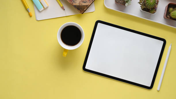 Top view image of colorful workplace. Pencil compass, coffee cup, notebook, marker pens, cactus, stylus pen and computer tablet with white empty screen flat lay on yellow table.