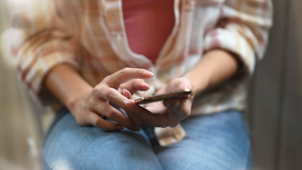 istock Cropped image of beautiful woman using/holding smartphone in her hands while sitting at the working desk over modern living room as background. 1216089676