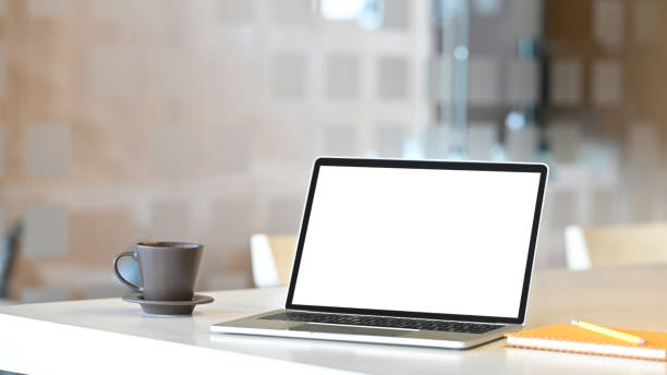 istock Photo of white blank screen computer laptop putting on white working desk with coffee cup, pencil and notebook over modern office as background. Orderly workplace concept. Space for advertisement. 1216089752