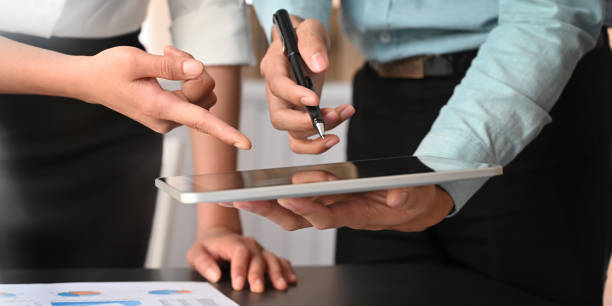 istock Cropped image of data analyst team working together with computer tablet and graphic charts information while standing together at modern meeting desk over meeting room as background. 1216090054