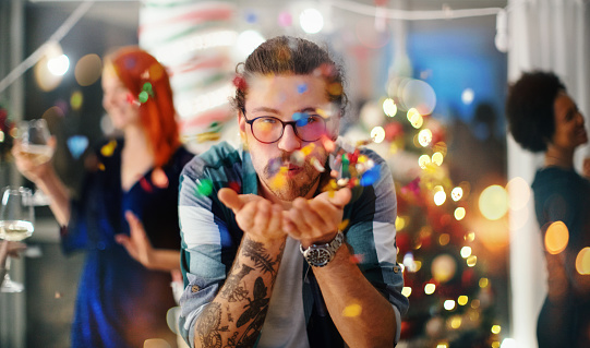 Friends celebrating Christmas together. Young man standing and blowing confetti in the air, looking at camera.