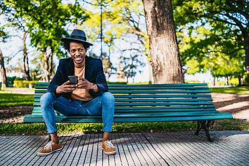 Smiling young African man using smartphone on park bench.