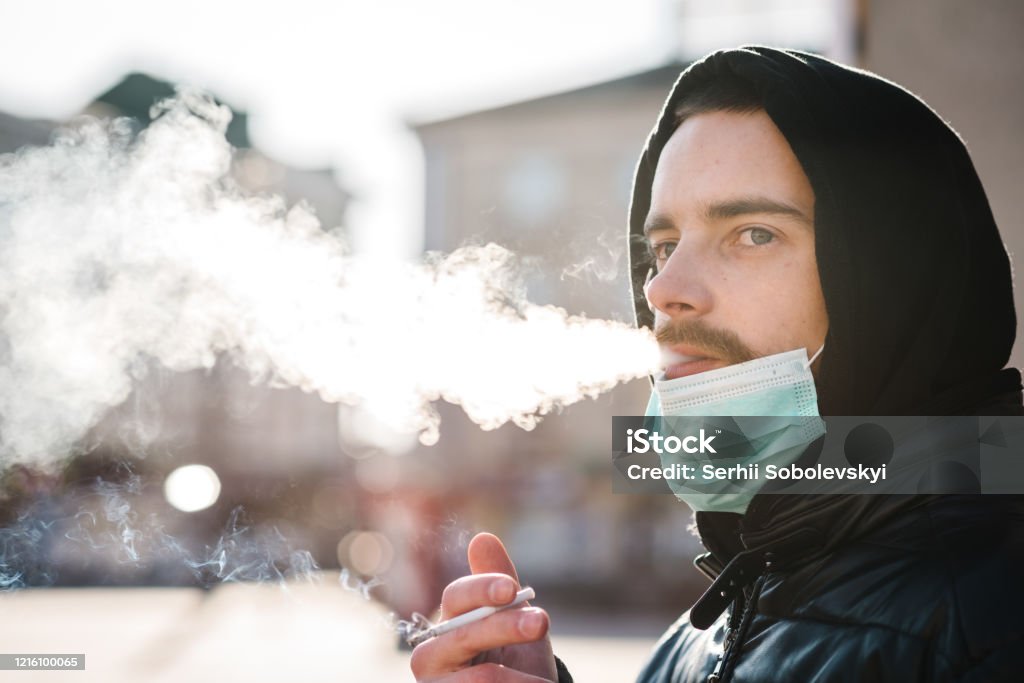 Rauchen. Nahaufnahme Mann mit Maske während COVID-19 Pandemie Rauchen einer Zigarette auf der Straße. Rauchen verursacht Lungenkrebs und andere Krankheiten. Die Gefahren und Schäden des Rauchens. Coronavirus. - Lizenzfrei Ansteckende Krankheit Stock-Foto