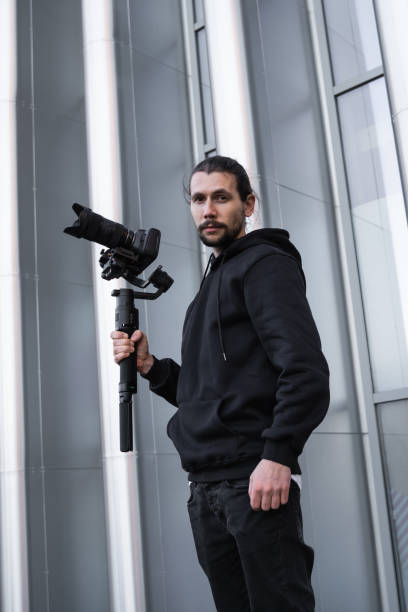 istock Young Professional videographer holding professional camera on 3-axis gimbal stabilizer. Pro equipment helps to make high quality video without shaking. Cameraman wearing white hoodie making a videos. 1216103810