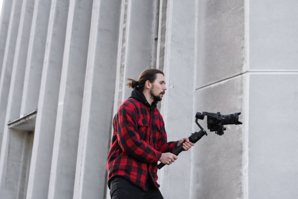 istock Young Professional videographer holding professional camera on 3-axis gimbal stabilizer. Pro equipment helps to make high quality video without shaking. Cameraman wearing red shirt making a videos. 1216103889