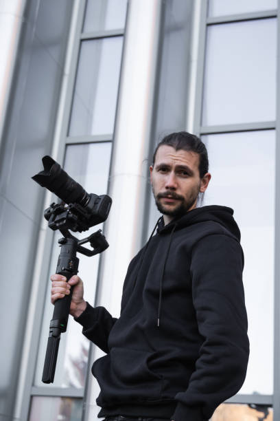 istock Young Professional videographer holding professional camera on 3-axis gimbal stabilizer. Pro equipment helps to make high quality video without shaking. Cameraman wearing white hoodie making a videos. 1216103957