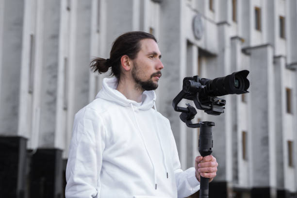 istock Young Professional videographer holding professional camera on 3-axis gimbal stabilizer. Pro equipment helps to make high quality video without shaking. Cameraman wearing white hoodie making a videos. 1216104073