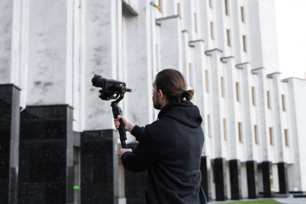 istock Young Professional videographer holding professional camera on 3-axis gimbal stabilizer. Pro equipment helps to make high quality video without shaking. Cameraman wearing black hoodie making a videos. 1216104187