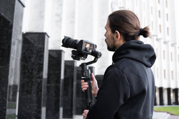 istock Young Professional videographer holding professional camera on 3-axis gimbal stabilizer. Pro equipment helps to make high quality video without shaking. Cameraman wearing black hoodie making a videos. 1216105040