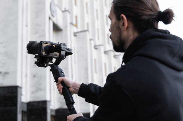istock Young Professional videographer holding professional camera on 3-axis gimbal stabilizer. Pro equipment helps to make high quality video without shaking. Cameraman wearing black hoodie making a videos. 1216105109