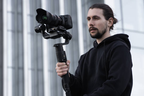 istock Young Professional videographer holding professional camera on 3-axis gimbal stabilizer. Pro equipment helps to make high quality video without shaking. Cameraman wearing white hoodie making a videos. 1216105188