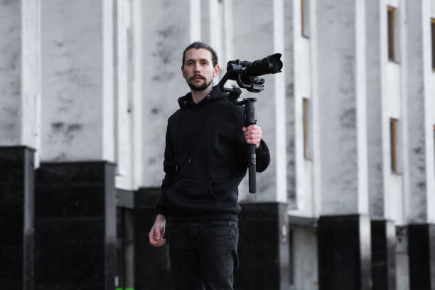 istock Young Professional videographer holding professional camera on 3-axis gimbal stabilizer. Pro equipment helps to make high quality video without shaking. Cameraman wearing black hoodie making a videos. 1216105403