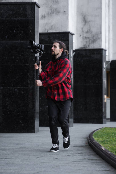 istock Young Professional videographer holding professional camera on 3-axis gimbal stabilizer. Pro equipment helps to make high quality video without shaking. Cameraman wearing red shirt making a videos. 1216105490
