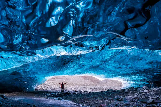 Inside a blue glacial ice cave in the glacier. Breioarmerkurjokull, part of the Vatnajokull glacier in southeast Iceland. A young woman stands in the cave opening to demonstrate scale.