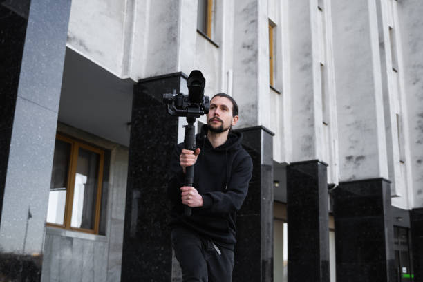 istock Young Professional videographer holding professional camera on 3-axis gimbal stabilizer. Pro equipment helps to make high quality video without shaking. Cameraman wearing black hoodie making a videos. 1216105723